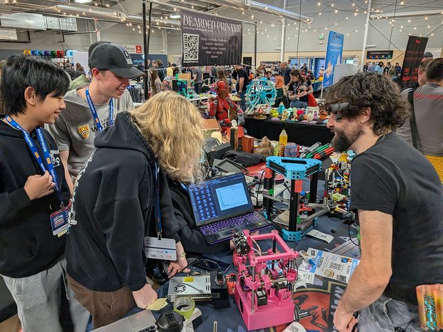 Teenagers talking with a visitor across a booth table covered in 3D printers at a maker convention.