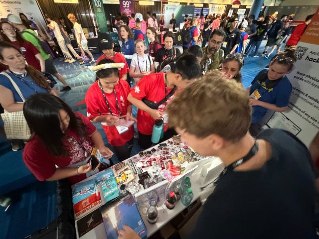 A crowded Hack Club booth indoors, people leaning over a table covered in stickers and giveaways.