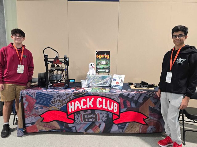 Two teenagers standing either side of a Hack Club booth table with a 3D printer, stickers and a printed tablecloth.